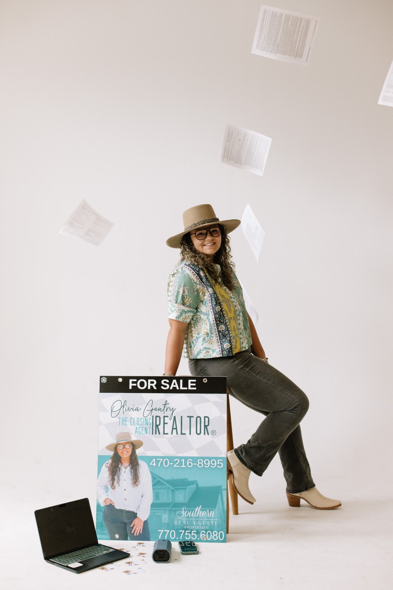 Olivia Gentry Williams seated beside her Southern Real Estate Properties For Sale sign as contract pages fly through the air